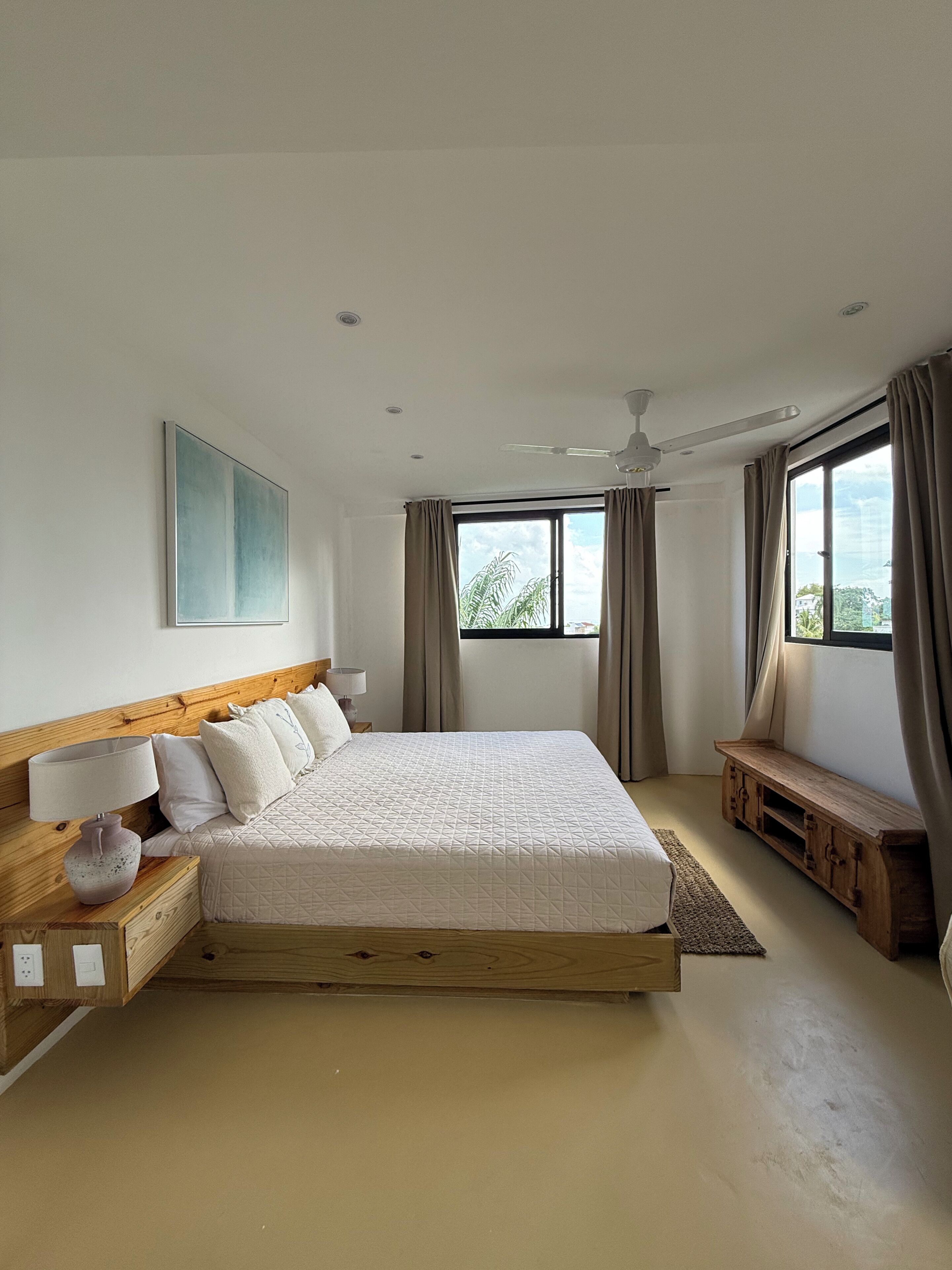 Un dormitorio con una cama grande, cabecera de madera, mesita de noche con lámpara, un banco y vistas por la ventana.
