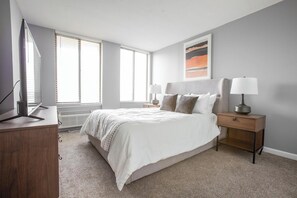 A modern bedroom with a bed, bedside tables, a TV, and a window with blinds. - Landing | Stunning 2BD, Pool, Gym (Fort Wayne)