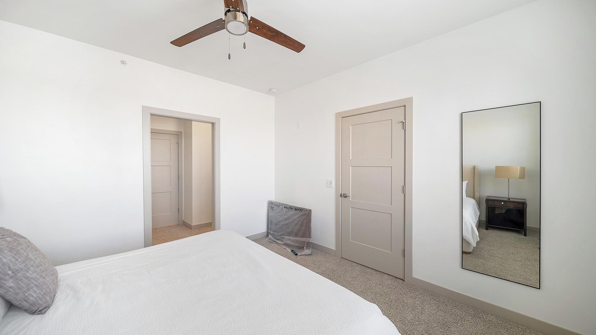 Un dormitorio con cama, ventilador de techo, puerta, espejo y mesita de noche con lámpara.
