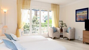 Habitación de hotel con una cama grande, dos sillones, un televisor y un balcón con cortinas amarillas.
