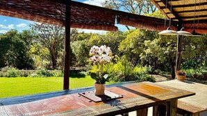 Un patio exterior con mesa y sillas de madera, un ventilador de techo y vistas a un jardín con árboles y césped.