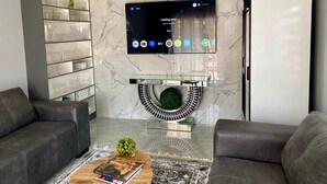 A modern living room with a flat-screen TV mounted on the wall, a marble-patterned wall, a glass coffee table, and two grey sofas. - Executive studio apartment near the airport - 2320 (Harare)