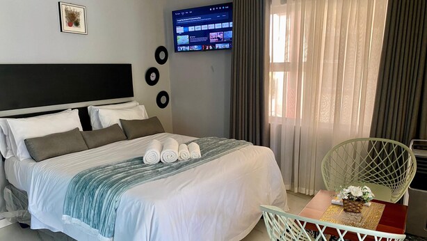 Un lit bien fait, agrémenté de serviettes pliées, un téléviseur fixé au mur diffusant un service de streaming, un élément décoratif mural, et un coin repas avec une chaise et une table.