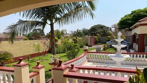 Un balcon doté d’un sol en pierre, de balustrades blanches, d’une fontaine blanche et d’un jardin agrémenté de palmiers et de fleurs rouges.