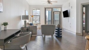 A modern living room with a sofa, chairs, a coffee table, a TV, and a balcony with large windows. - Landing | Incredible 1BD, Clubhouse, Gym (Atlanta)