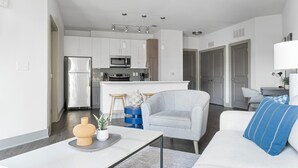 A modern living room with a grey sofa, a coffee table, and a blue pillow. There is a kitchen area with white cabinets, a refrigerator, and a microwave. - Landing | Incredible 1BD, Clubhouse, Gym (Atlanta)