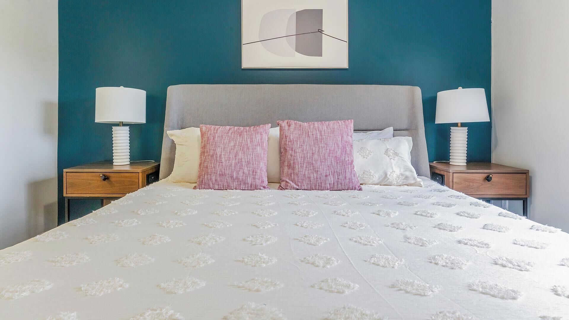 A neatly made bed with a white duvet and patterned bedding, flanked by two bedside tables with lamps, against a teal wall with a simple abstract artwork above.