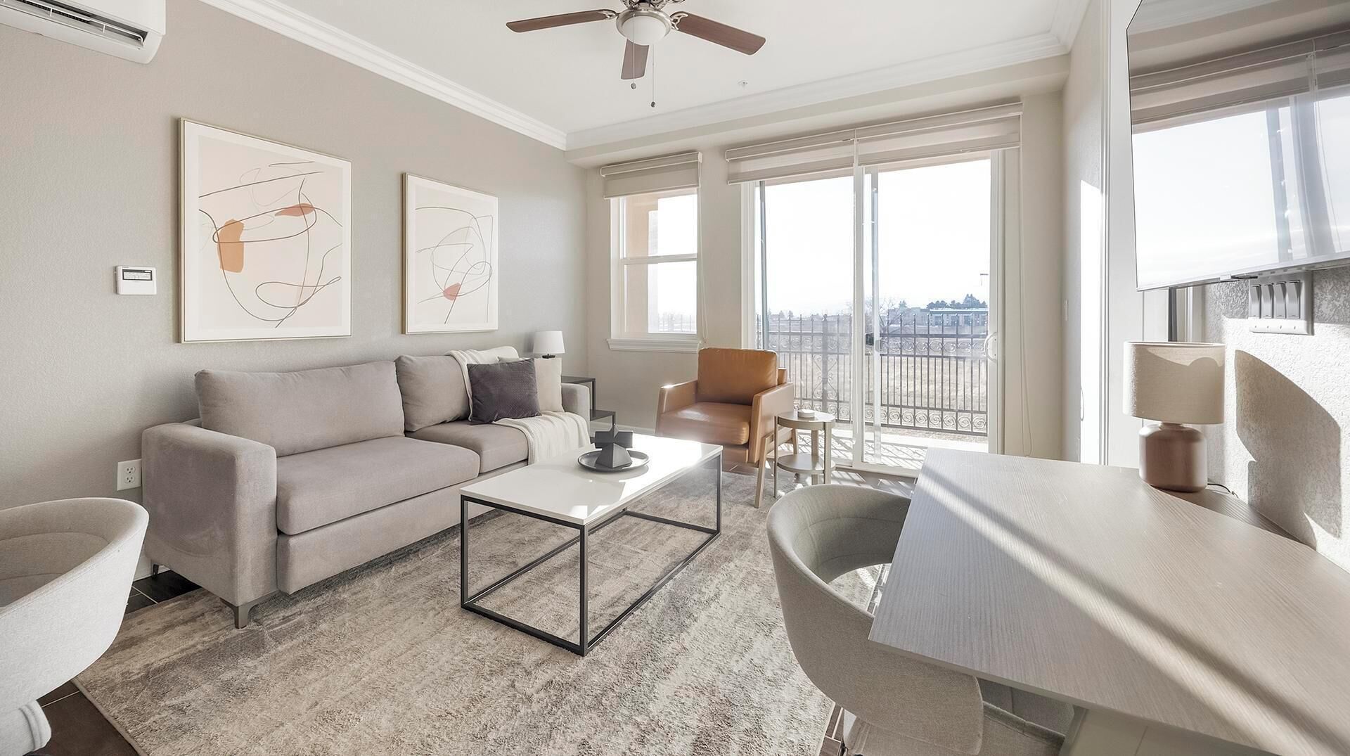 Una sala de estar moderna con un sofá, una mesa de centro y un sillón. Hay dos ventanas amplias que dan a una vista de la ciudad.