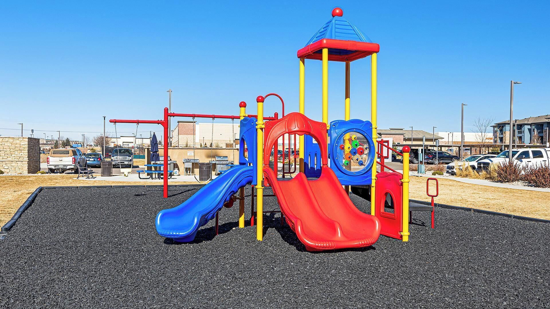 A playground with two slides and a climbing structure, surrounded by a parking lot and buildings.