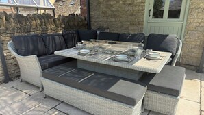 A wicker outdoor seating area with cushions, a table set for dining, and a stone wall backdrop. - Granary Cottage (Chippenham)