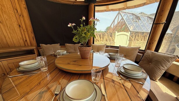 A round wooden dining table with plates, cutlery, and glasses, surrounded by cushioned chairs, with a potted plant and a view of a building through a window. - Granary Cottage (Chippenham)