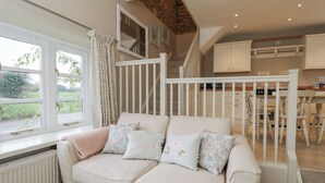A cozy room with a white sofa, a window with curtains, a staircase, and a kitchen area. - Granary Cottage (Chippenham)