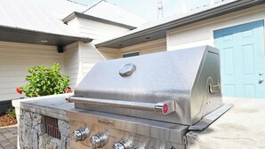 A stainless steel outdoor grill with four control knobs, placed on a stone base, in front of a house with a blue door and a small garden with red flowers. - Landing | Stunning 1BD, Courtyard, Pool (Nashville)