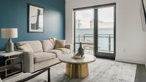 Een moderne woonkamer met een bank, een salontafel en een lamp. Er is een groot raam met uitzicht op gebouwen en een balkon.