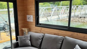 Ein modernes Blockhaushaus-Interieur mit einem grauen Ecksofa, Holzwänden und großen Fenstern mit Blick auf eine Brücke und viel Grün.