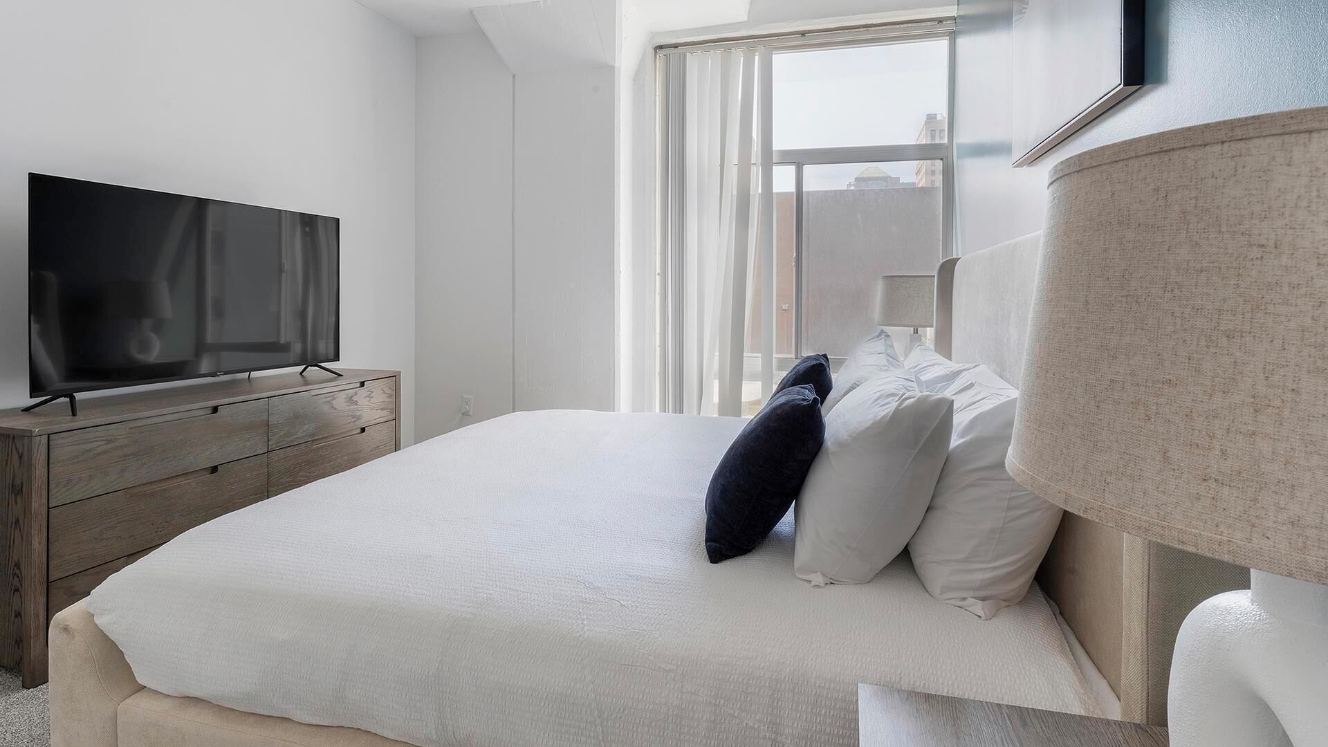A modern hotel room with a large bed, a flat-screen TV on a wooden dresser, a window with sheer curtains, and a lamp on the bedside table.