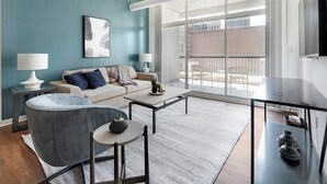Un salon moderne avec un canapé, une table ronde, un fauteuil et une grande fenêtre donnant sur des immeubles.