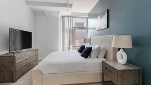 Une chambre d’hôtel moderne avec un grand lit, un téléviseur posé sur une commode en bois, une lampe de chevet et une vue sur la ville par la fenêtre.
