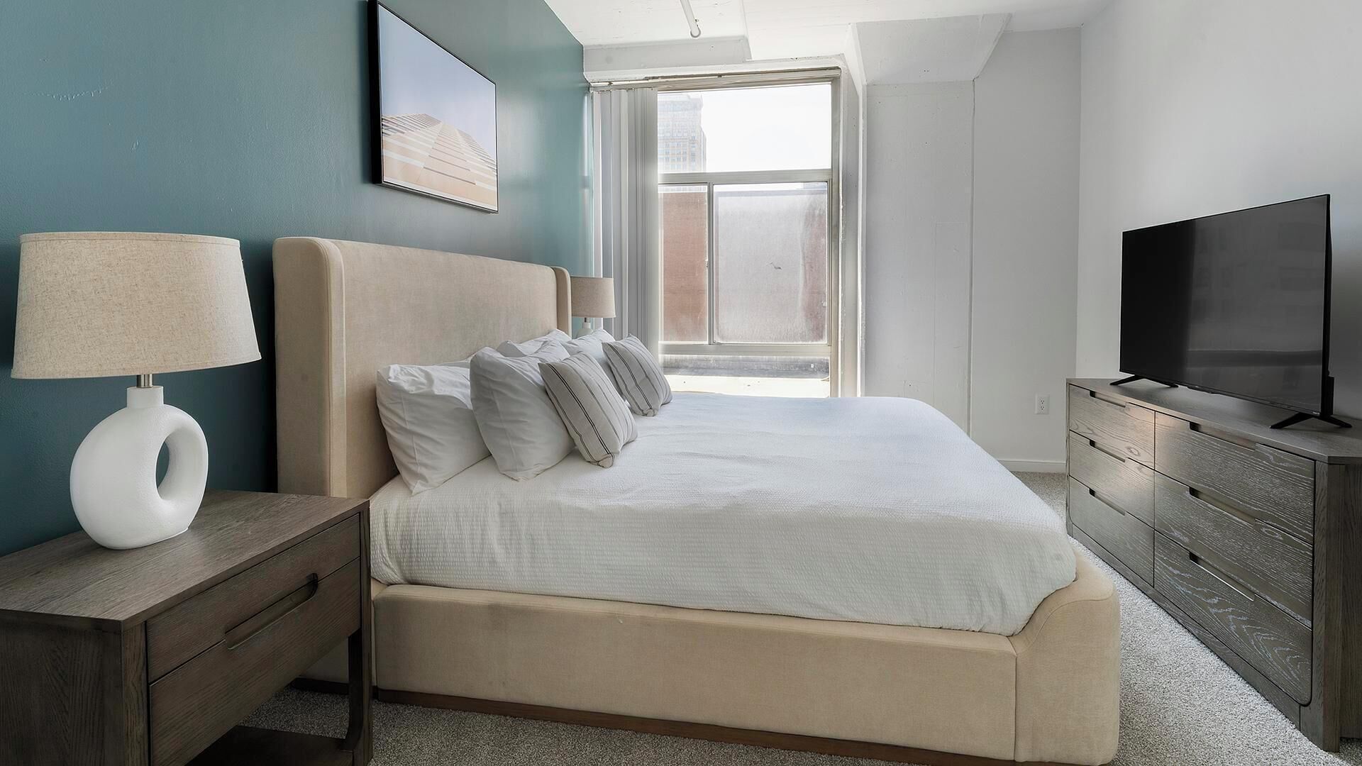 A modern hotel room with a large bed, a bedside table with a lamp, a TV, and a window with a view of the city.
