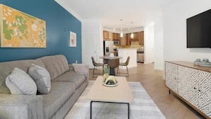 Een moderne woonkamer met een grijze bank, een salontafel, en een eethoek met een ronde tafel en stoelen. Achtergrond is een keuken met houten kastjes en een flatscreen televisie aan de muur bevestigd.