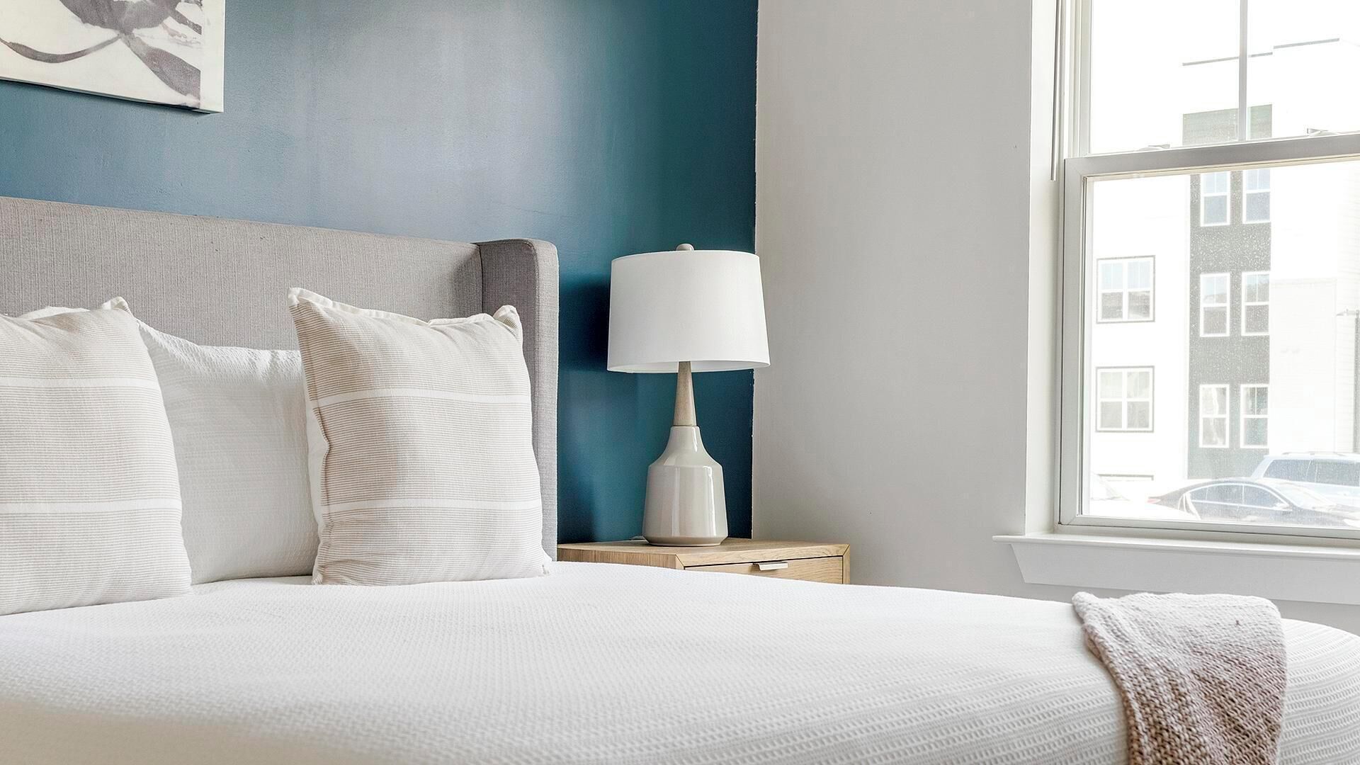Een opgemaakt bed met kussens, een bedlampje en een raam met uitzicht op gebouwen.