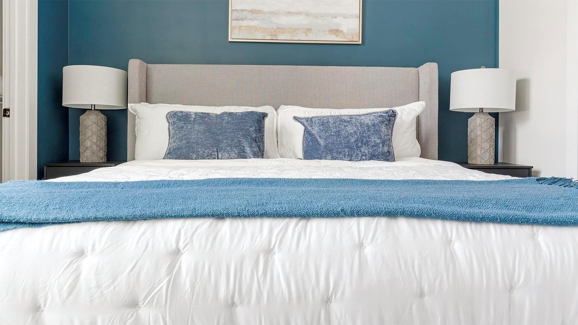 A neatly made bed with a white duvet and blue throw blanket, flanked by two bedside tables with lamps, against a blue wall with a framed artwork above.