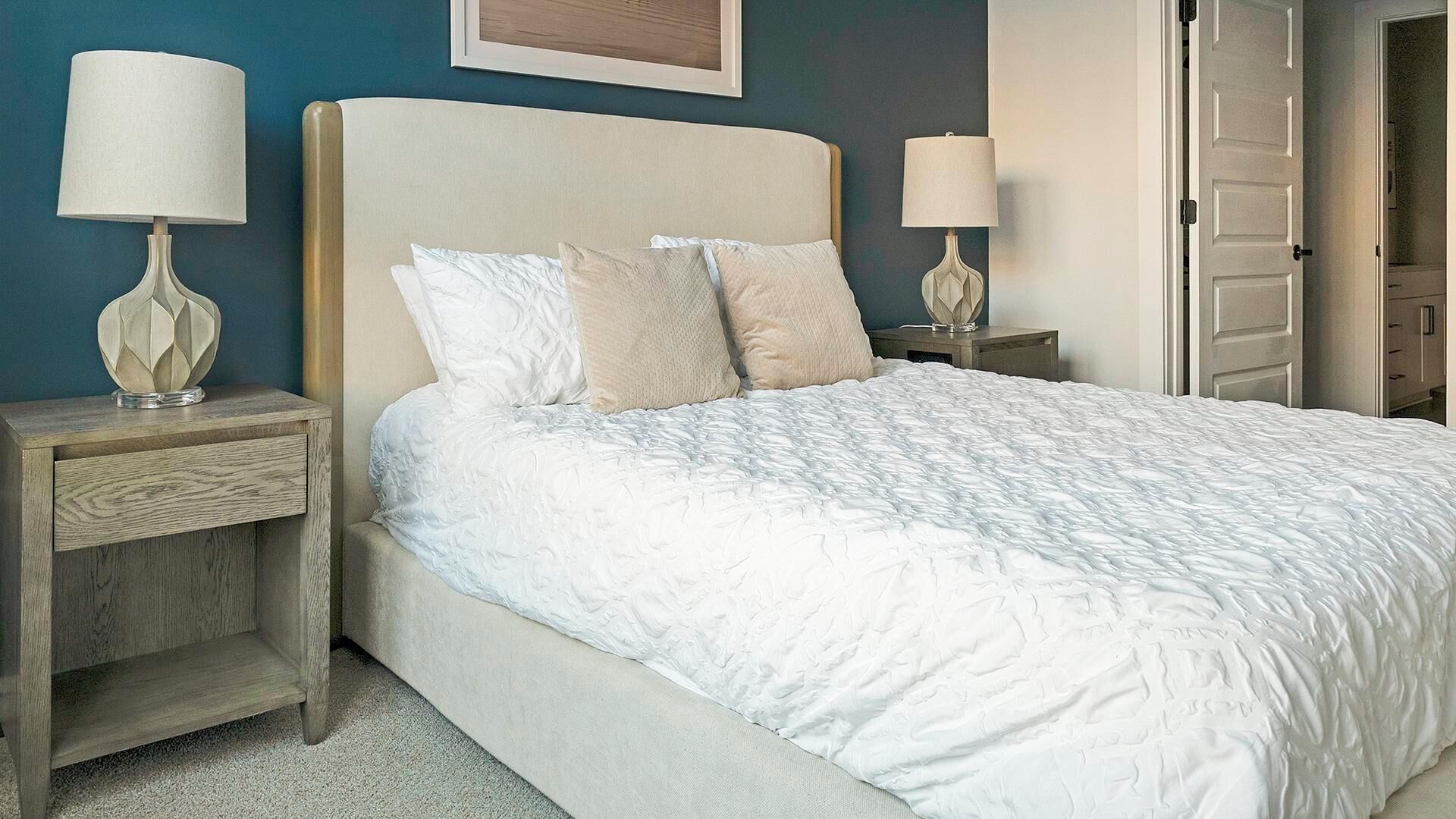 Una cama bien hecha con edredón blanco y almohadas, dos lámparas de noche, una mesita de madera y una puerta a la derecha.