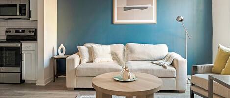 A living room with a beige sofa, a round coffee table, a floor lamp, and a kitchen area with appliances.