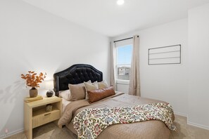Une chambre à coucher avec une tête de lit capitonnée, une couvre-lit à motifs floraux, une table de chevet avec une lampe et une fenêtre avec des stores.