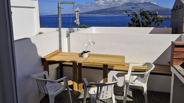 A balcony with a wooden table and white plastic chairs, overlooking a coastal view with a mountain in the background.