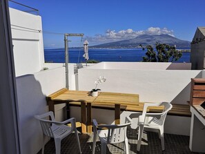 Ein Balkon mit Holztisch und weißen Plastikstühlen, der Blick geht auf die Küste mit einem Berg im Hintergrund.