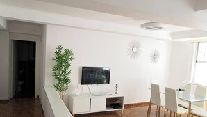 A modern living room with a flat-screen TV, white console, dining table with chairs, and a potted plant.