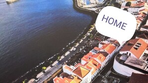 An aerial view of a coastal town with a prominent waterfront promenade, a large white building with orange roofs, and a speech bubble with the word "HOME".