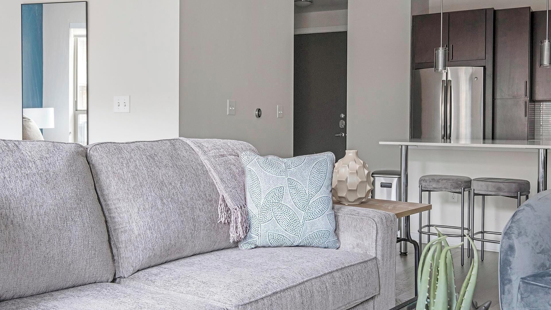 En moderne stue med en grå sofa, et mønstret pude og et minimalistisk køkkenområde med rustfrie stålens hvidevarer.