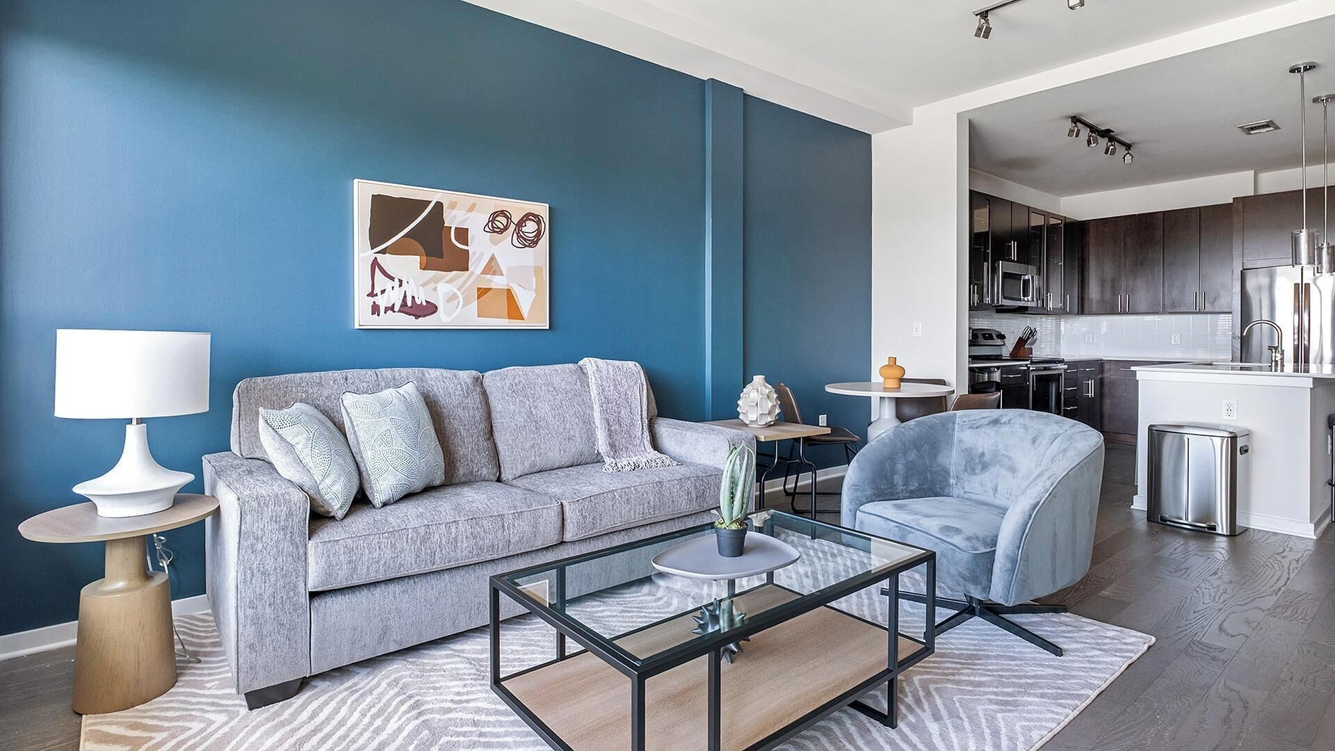 Una sala de estar moderna con un sofá gris, un sillón azul, una mesa de centro de vidrio y una cocina al fondo.