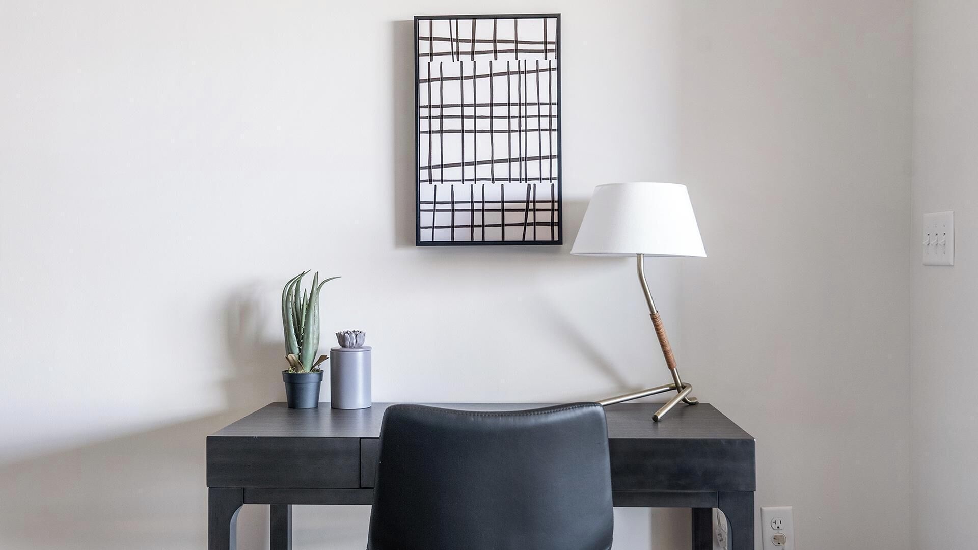 A modern desk with a lamp, a framed artwork, and two potted plants.