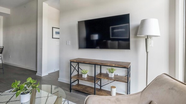 A modern living room with a large flat-screen TV mounted on the wall, a wooden console table with shelves, a floor lamp, and a beige sofa.