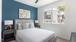 A bedroom with a bed, two bedside tables with lamps, a large window with a view of buildings, and a wall with abstract art.