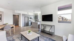 A modern apartment with a dining area, kitchen, living room, and a large window offering a city view.