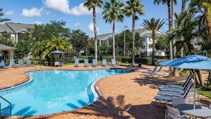 A swimming pool with lounge chairs and umbrellas, surrounded by palm trees and buildings. - Landing | Chic 2BD, Gym, Pool (Orlando)