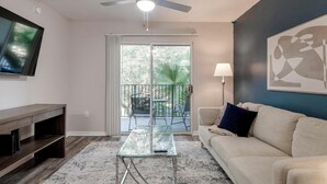 A living room with a glass coffee table, a sofa, a TV mounted on the wall, and a balcony with a view of trees and a palm plant. - Landing | Chic 2BD, Gym, Pool (Orlando)
