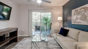 A living room with a glass coffee table, a sofa, a TV mounted on the wall, and a balcony with a view of trees and a palm plant. - Landing | Chic 2BD, Gym, Pool (Orlando)