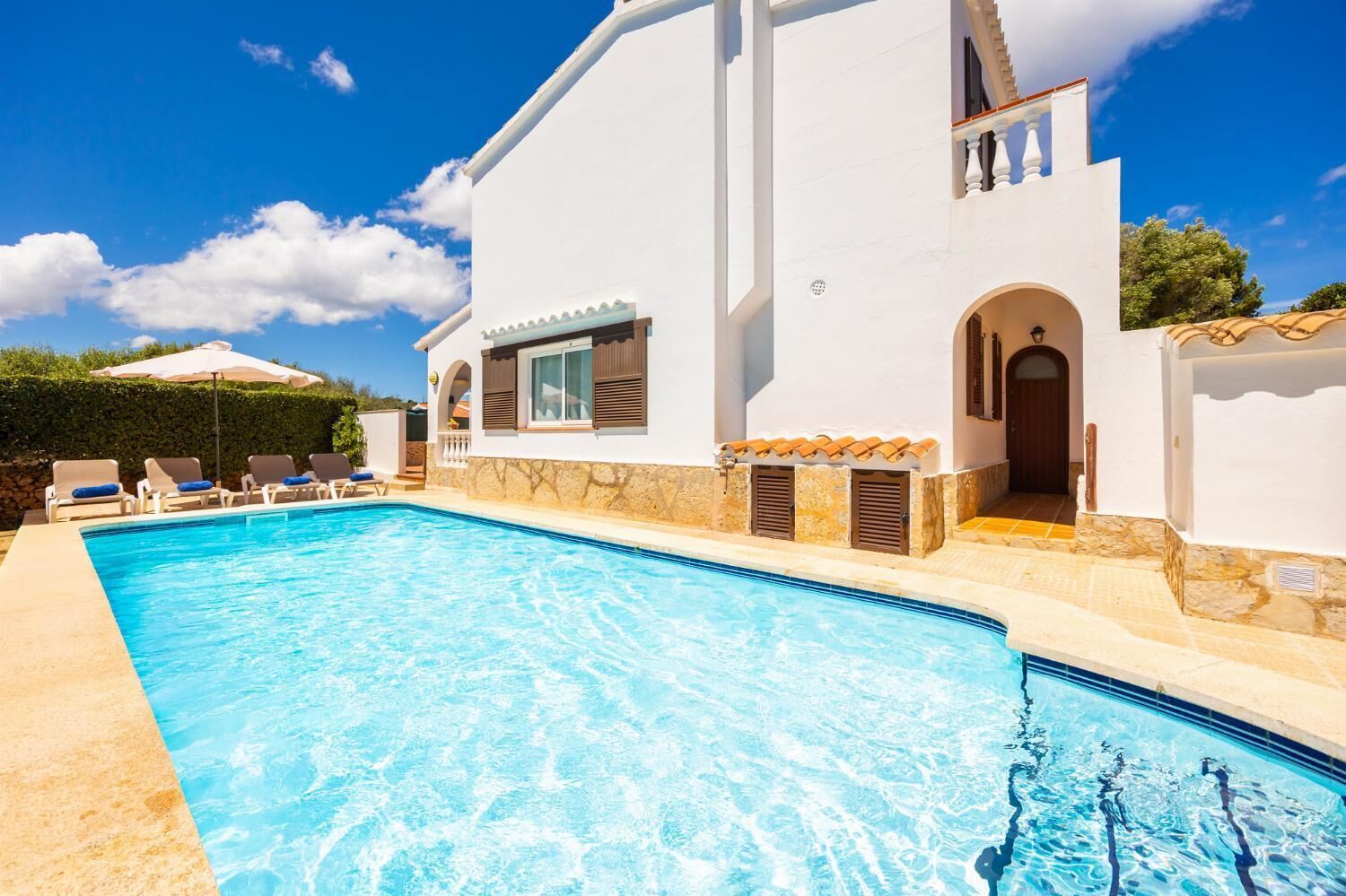 A swimming pool adjacent to a white building with a stone facade, a covered patio, and a clear blue sky.