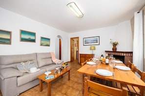 Un salon comprenant un canapé, une table basse et une table à manger pour quatre personnes. Les murs sont ornés de tableaux encadrés, il y a une cheminée et une porte qui donne sur une autre pièce.
