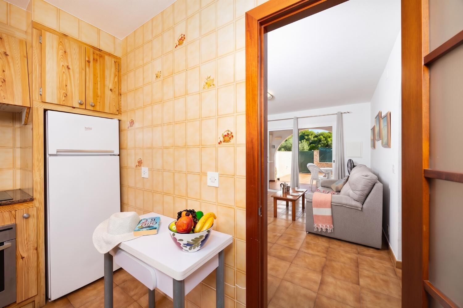 Une cuisine de taille réduite, avec des meubles en bois, un réfrigérateur blanc et une table sur laquelle se trouve un saladier rempli de fruits.
