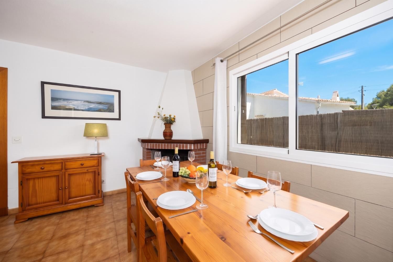 Uma sala de jantar com mesa de madeira posta para quatro pessoas, lareira, quadro na parede e uma janela grande.