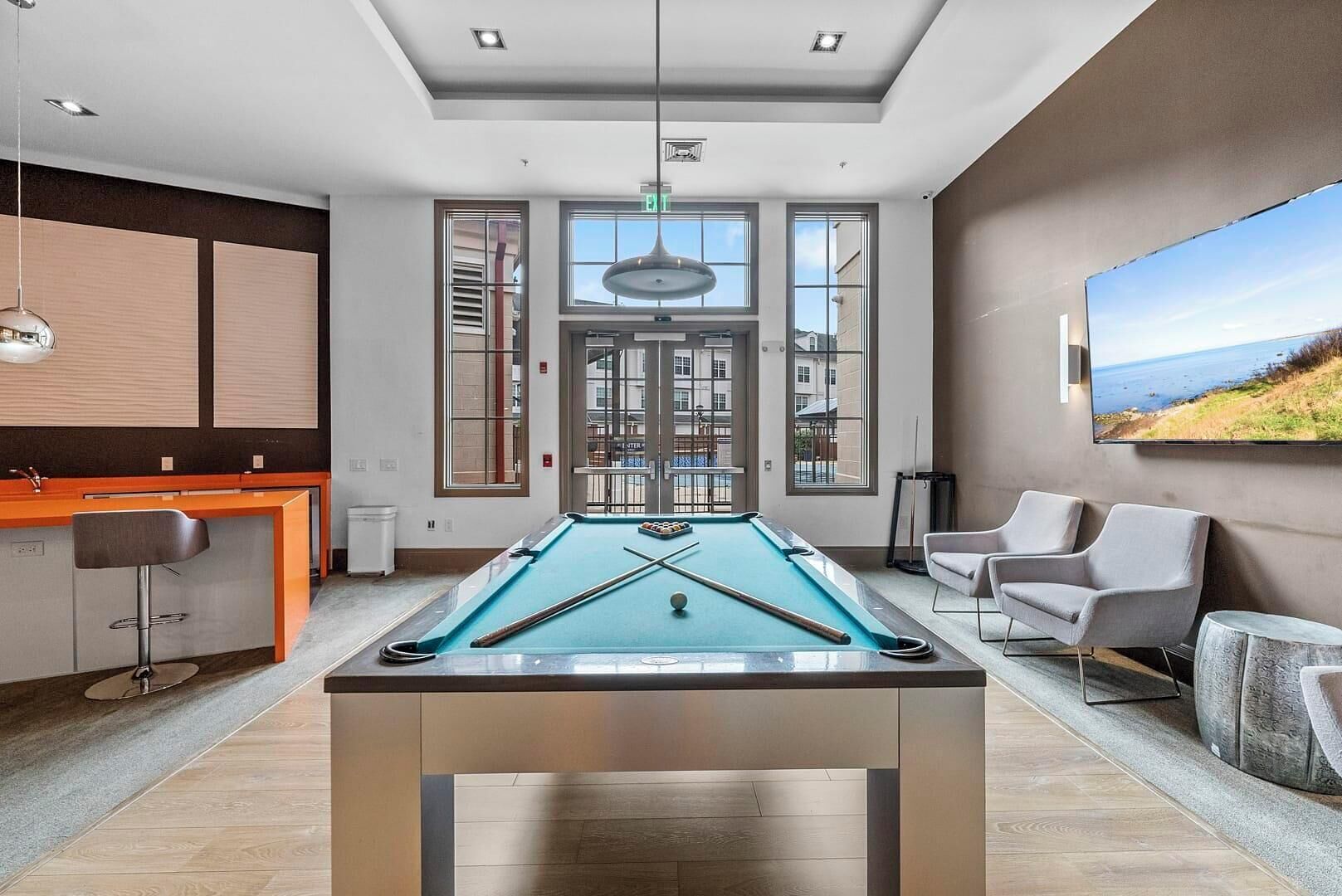 A pool table in a modern room with a bar area, seating, and large windows.