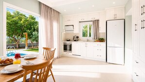 Una cucina moderna con mobili bianchi, un tavolo da pranzo con sedie, un frigorifero e vista su una piscina e vegetazione all'esterno.