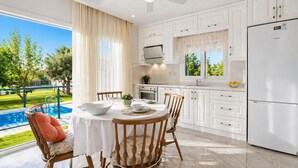 A modern kitchen with white cabinets, a dining table with chairs, a refrigerator, and a view of a swimming pool. - Villa Riva - Three Bedroom Villa, Sleeps 6 (Oludeniz)