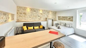 A modern room with a stone wall, a wooden table, a sofa with yellow cushions, a bed with striped bedding, and a large window. - 4★ Renovated Studio with Wifi, 5 min Walk to Quimper Station (Quimper)